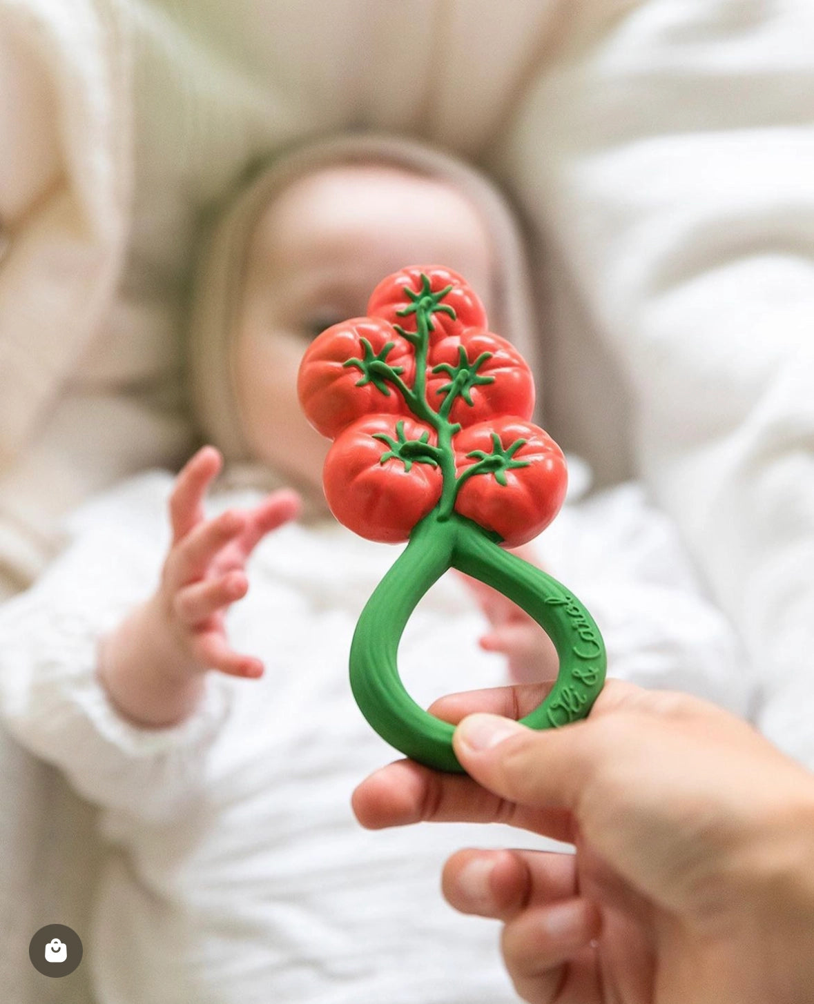 Hand holding a natural rubber tomato-shaped baby rattle toy in front of an out-of-focus baby reaching for it