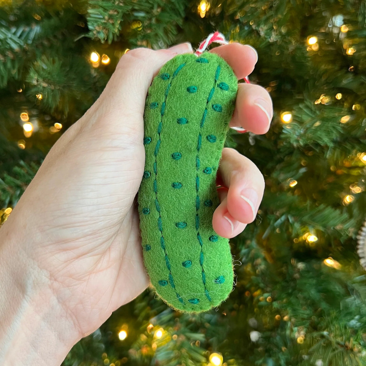 Hand holding a green Pickle Felt Ornament in front of a decorated Christmas tree with lights.