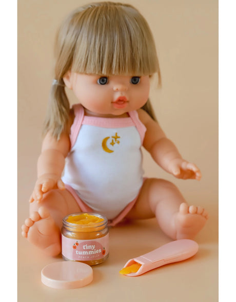 Doll sitting with peach Tiny Tummies food jar open and magic spoon with food on neutral background