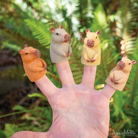 Hand wearing four colorful Finger Capybara soft vinyl finger puppets against a blurred green foliage background.