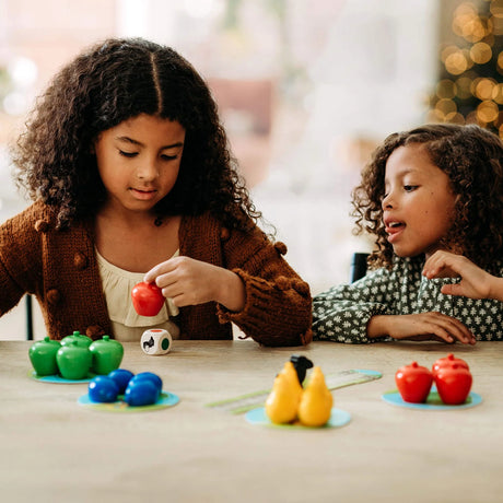 Two children playing with colorful wooden fruit pieces and a raven figure on a table in a cooperative game setting