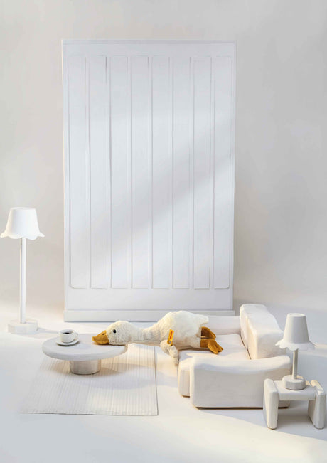 Plush Goose Gilda with long neck and yellow beak resting on a miniature white sofa in a cozy dollhouse setting.