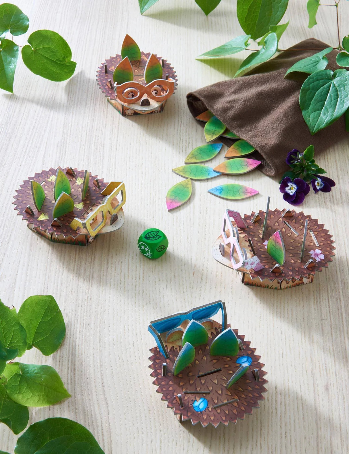 Four colorful hedgehog game pieces with leaves and dice on a wooden table surrounded by green plants.