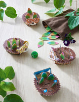 Four colorful hedgehog game pieces with leaves and dice on a wooden table surrounded by green plants.
