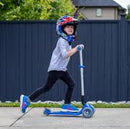 Helmeted child riding a blue scooter along a sidewalk beside a dark fence