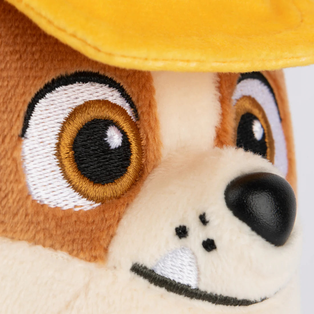 Close-up of a soft Plush toy dog wearing a yellow cap with embroidered eyes and nose, showing a friendly expression.