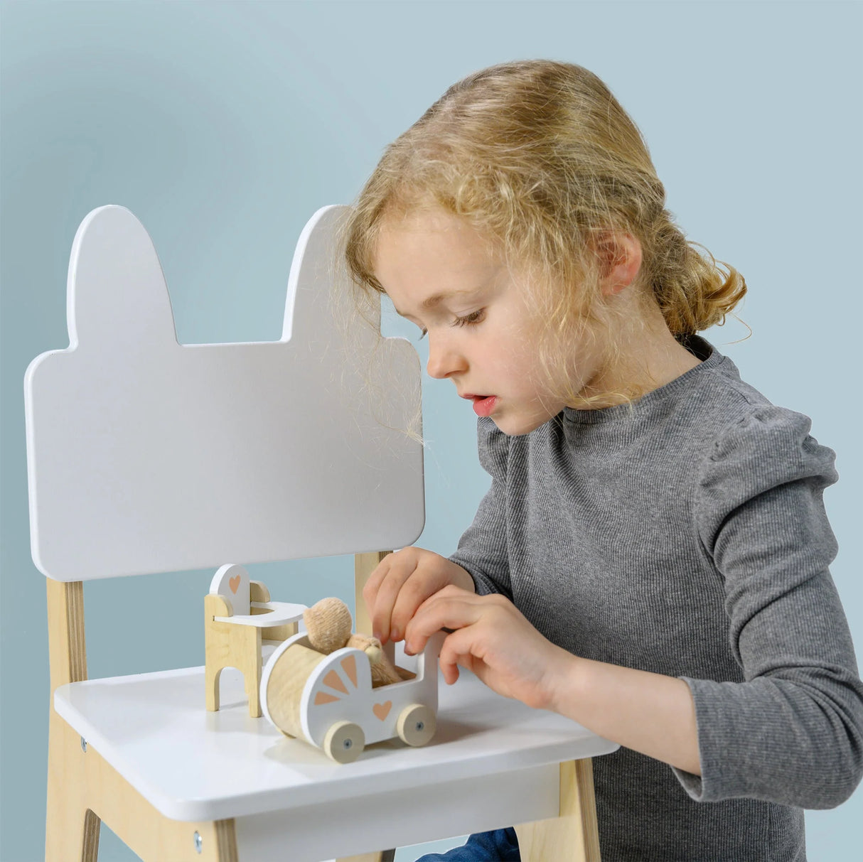 Young child playing with a wooden doll in a pram and highchair nursery set on a white chair