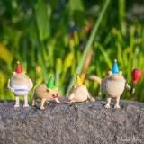Four colorful Party Frogs rain frog figurines in party hats, posed on a stone ledge with greenery background.