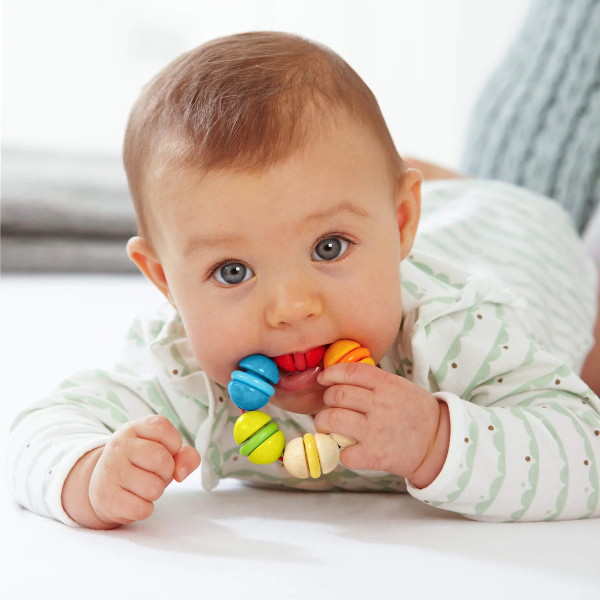 Baby lying down chewing on a colorful wooden rainbow rattle grasping and teething toy designed for sensory play