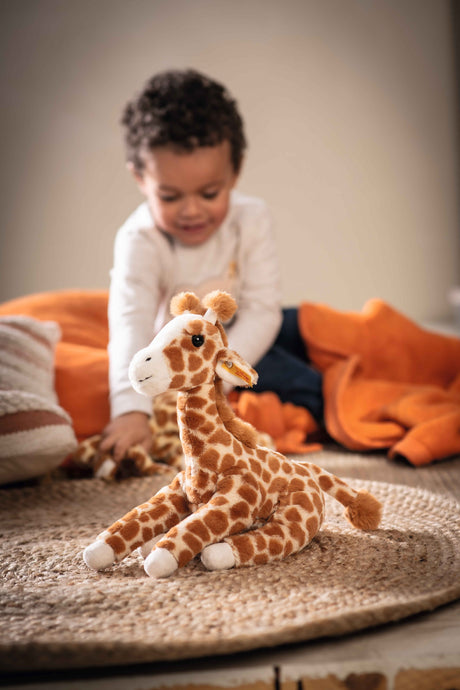 Plush giraffe stuffed animal with spotted fabric and fuzzy horns, named Giraffe Gina, sitting on a rug with child in background.