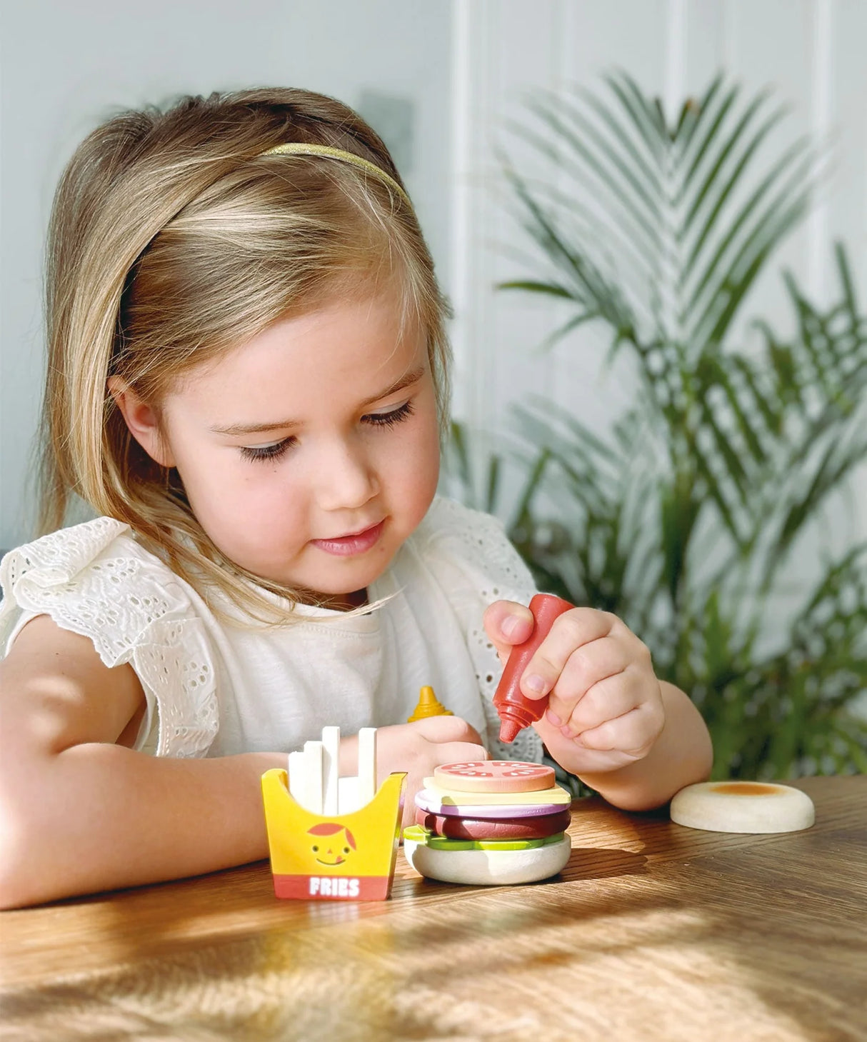 Young girl playing with a wooden take-out burger set stacking pretend burger ingredients at a wooden table