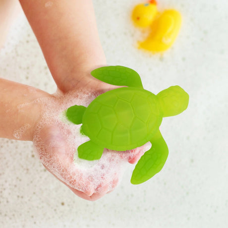Child holding a green Tub Turtle bath toy producing colorful light in soapy water with a yellow rubber duck in background