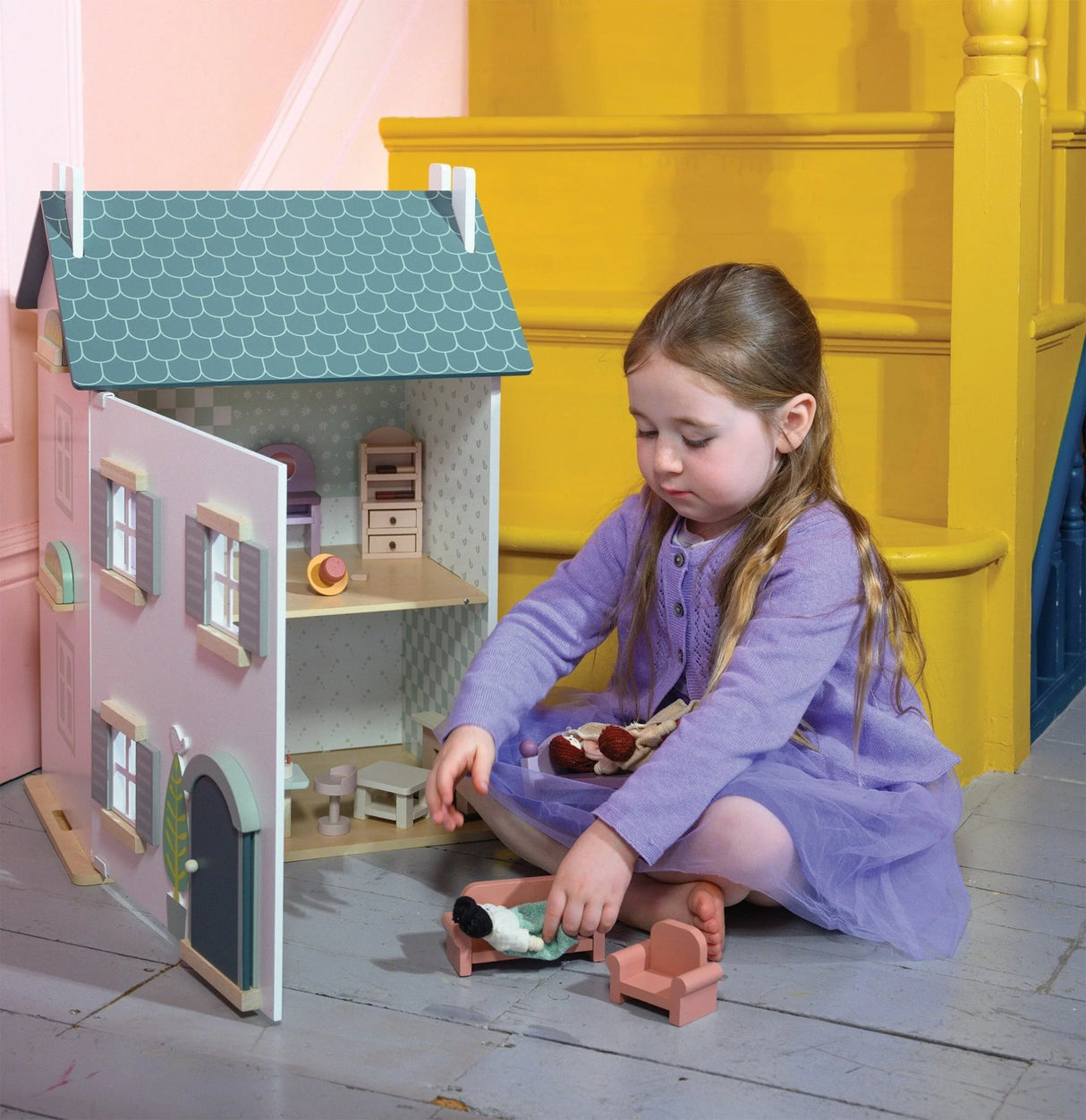 Young girl in purple dress playing with medium-sized wooden Willow Dollhouse on gray floor near yellow stairs