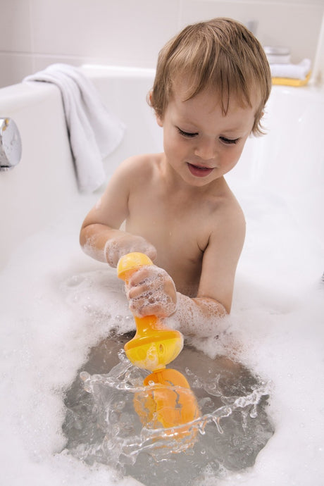 Child playing with yellow Bubble Bath Whisk in foam-filled bathtub creating bubbles and splashing water.