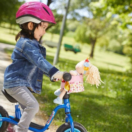 Child wearing pink helmet rides bike with Doll's Bicycle Seat holding a floral fabric doll securely on handlebars outdoors.