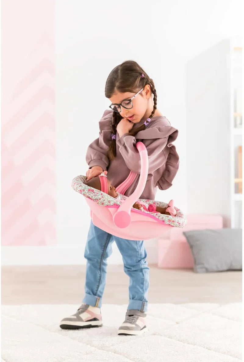 Young girl carrying a doll in a pink Car Seat Carrier Floral with flower print handle indoors.