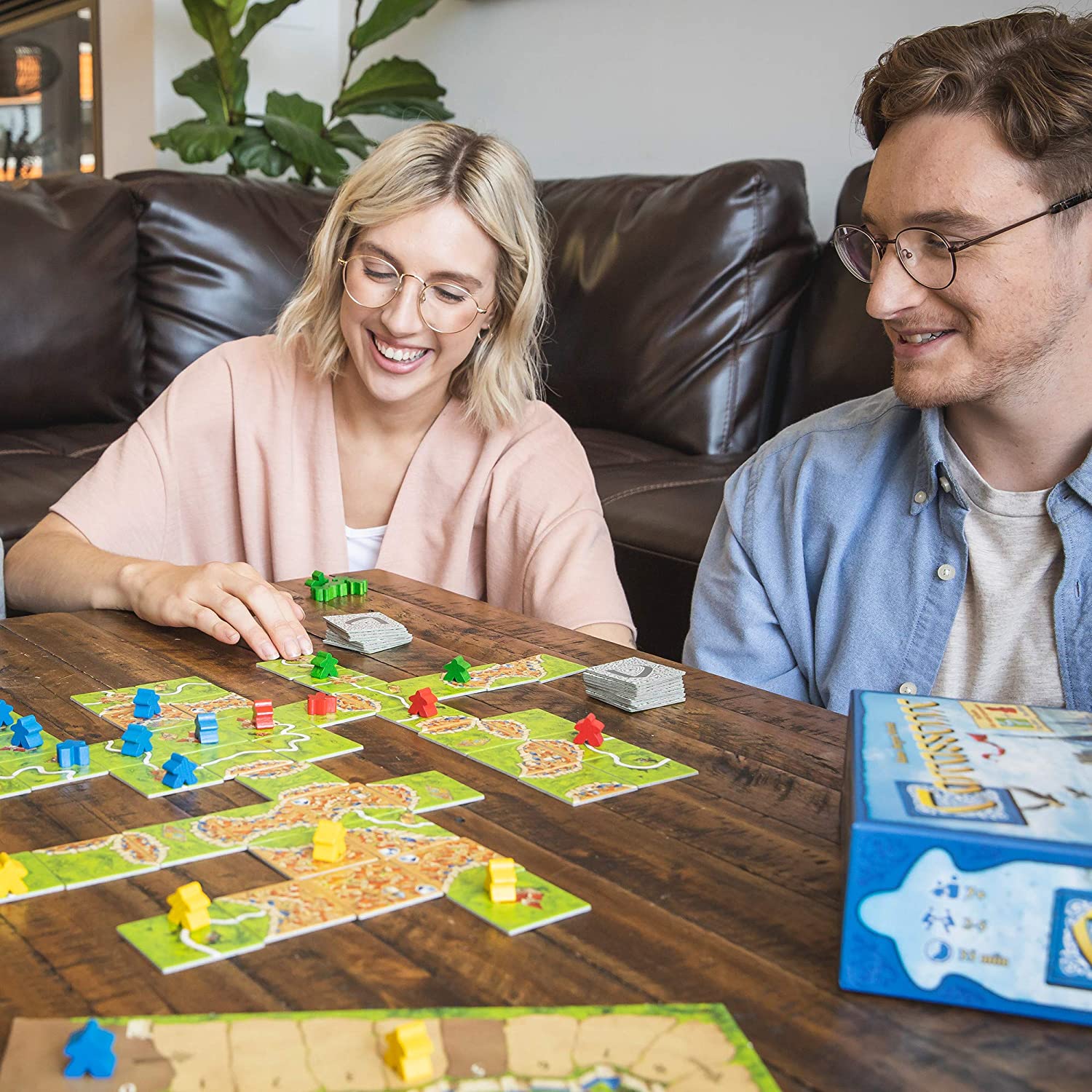 Two people smiling and playing Carcassonne tile-laying game on a wooden table with game pieces and box visible.