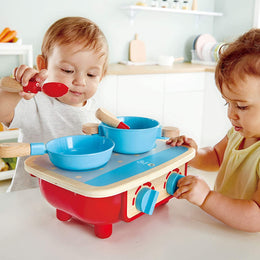 Two toddlers playing with a red and blue Kitchen Set wooden stove and cookware toy on a white table.