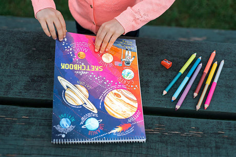 Solar System Sketchbook with colorful planets illustration, held by a child, alongside pencils and sharpener on a wooden table.