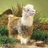 Folkmanis Alpaca Puppet with soft, woolly body and gentle expression standing among grass and rocks outdoors.
