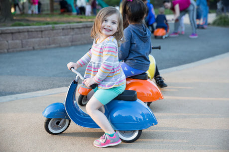 Child riding a blue self-propelled scooter toy Primo Ride On Blue in a park setting.