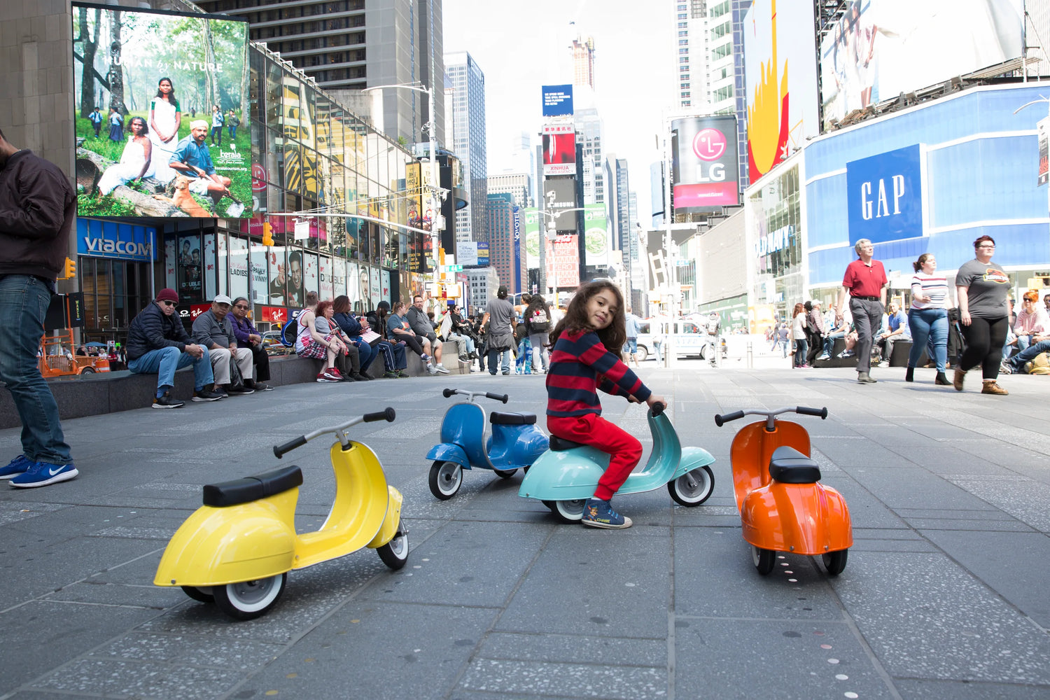 Primo Ride On Mint shown with yellow, blue, and orange ride-on scooters on city pavement with children playing nearby.