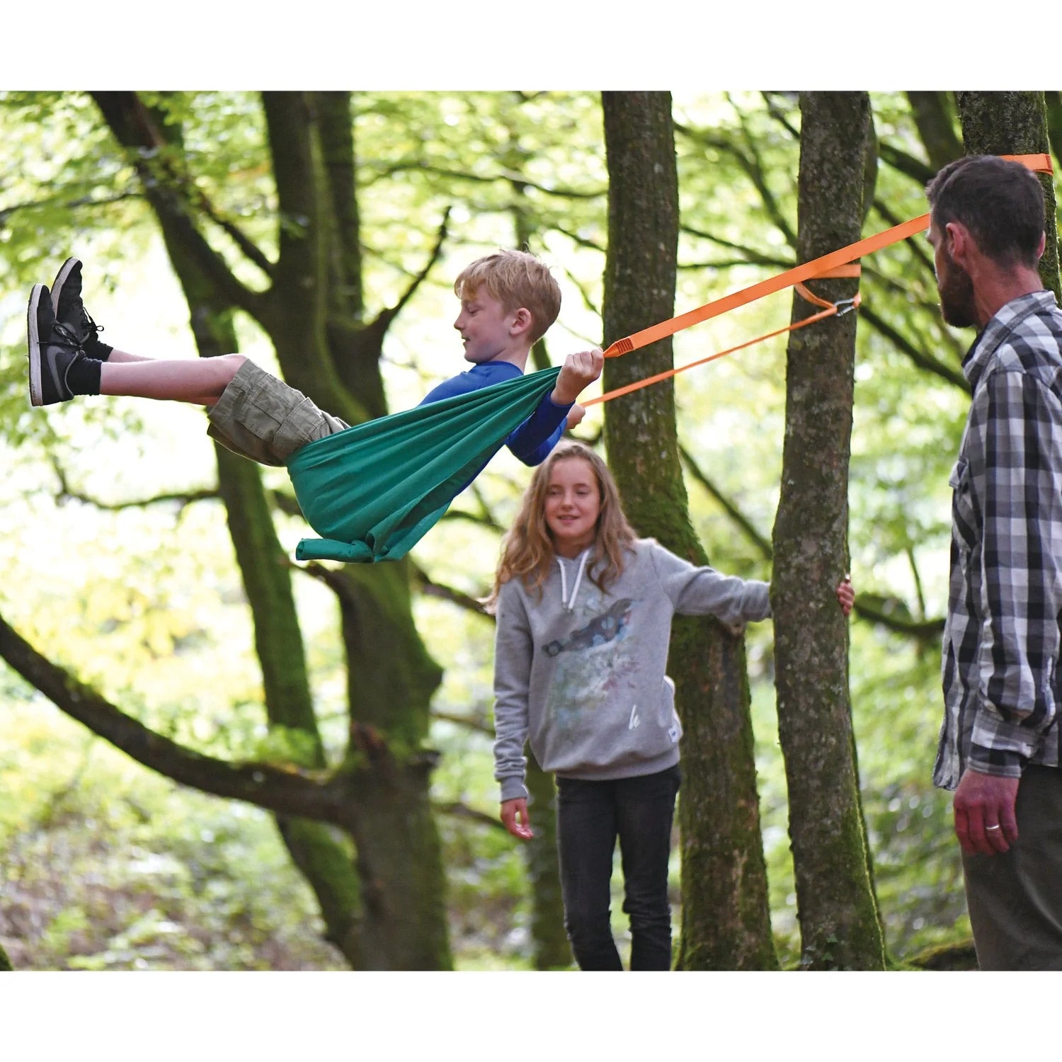 Child enjoying the Pocket Swing set up between two trees with adults in a green forest setting outdoors.