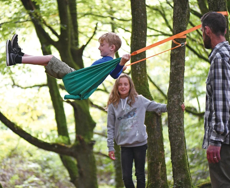 Child enjoying the Pocket Swing set up between two trees with adults in a green forest setting outdoors.