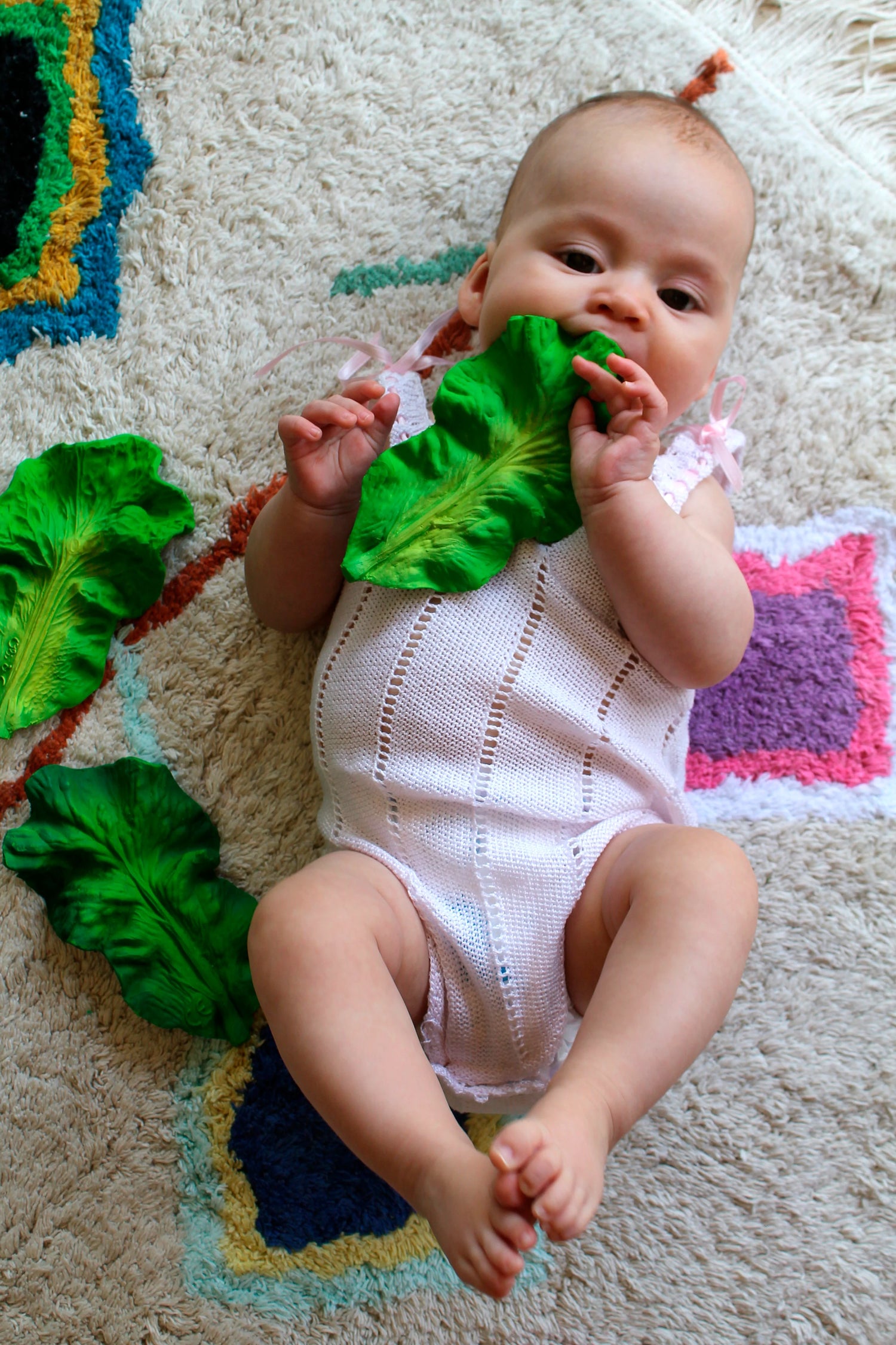 Baby chewing on a green vegetable-shaped teether lying on colorful rug, Natural Teether | Kendell the Kale.