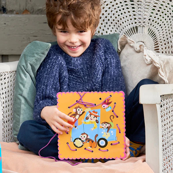 Child sitting on chair smiling and holding a colorful lacing card from Lacing Cards Good Deeds set with animal illustrations.