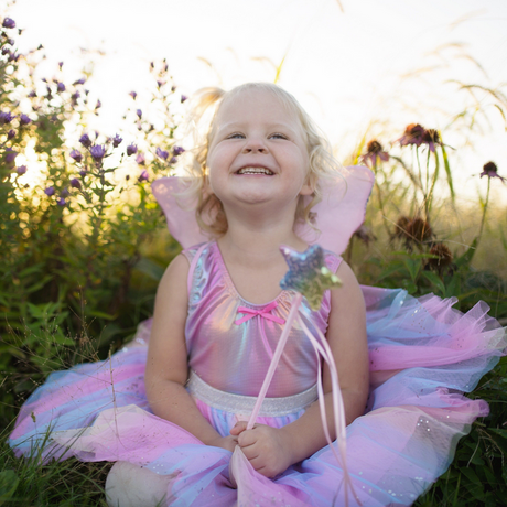 Smiling child wearing Rainbow Sequin Skirt, Wings & Wand set with pink wings and plush sequin wand outdoors.