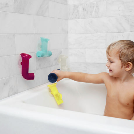 Child playing with colorful PIPES bath toys suctioned to tub wall, scooping and pouring water during bath time fun.