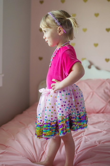 Child wearing Sequins Skirt | Party Fun Size Multi with rainbow sequins, standing on a pink bed in a decorated room.