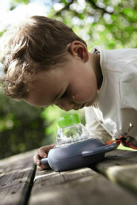 Young boy exploring nature using the Terra Kids | Exploration Magnifying Glass on a wooden surface outdoors.
