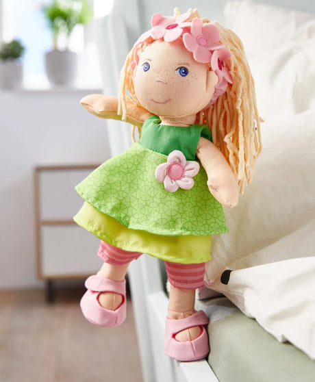 Soft Doll | Mali with embroidered face, floral headband, green dress, and pink shoes sitting on a bed edge.