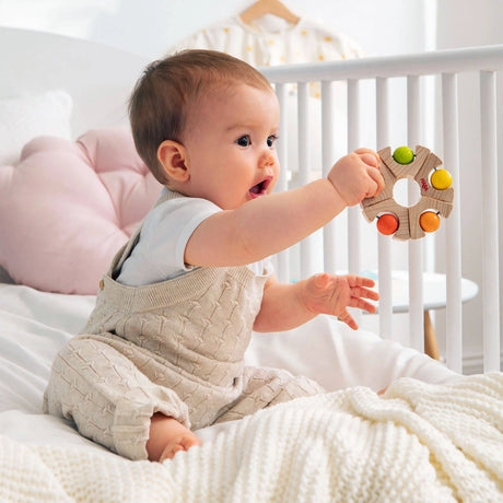 Baby sitting on bed playing with colorful Ball Wheel Grasping Toy with rattling balls for ages 6 months and up.
